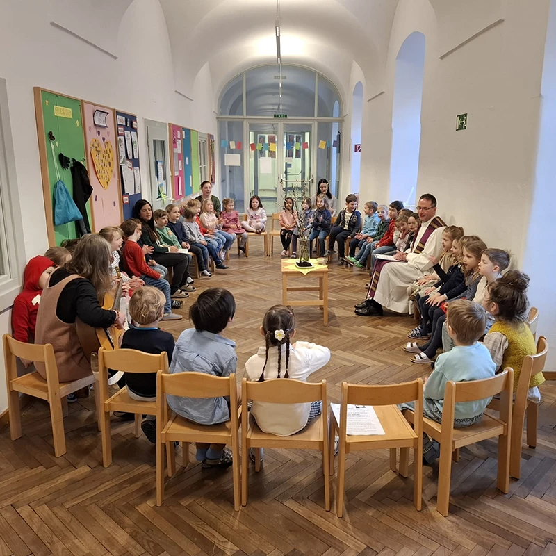 Die Kinder der Kindergruppe sitzen im Sesselkreis mit ihren Pädagoginnen und einem Pater zusammen. In der Mitte steht ein Tischchen mit einer Vase mit Palmzweigen.