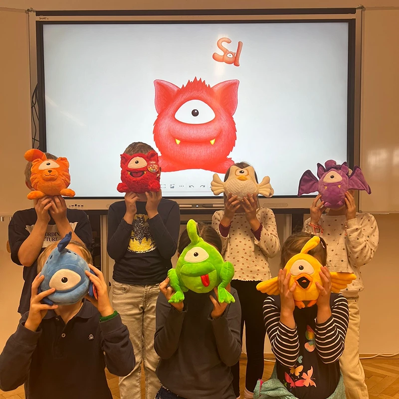 7 Kinder halten sich die Monsterfreunde-Kuscheltiere vor das Gesicht. Am Bild im Hintergrund ist der Monsterfreund Sol zu sehen.